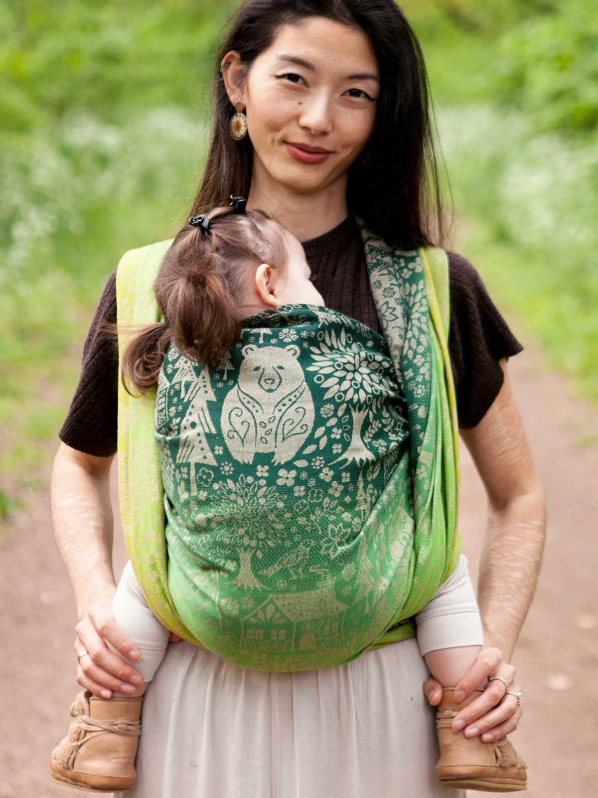 Woman carrying a baby in a green woven wrap with animal folk design.