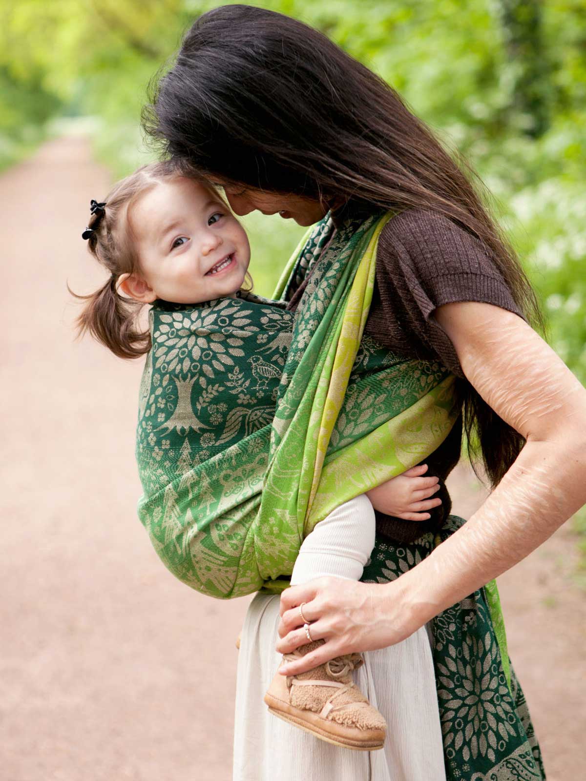 Woman carrying a baby in a green woven wrap with animal folk design.