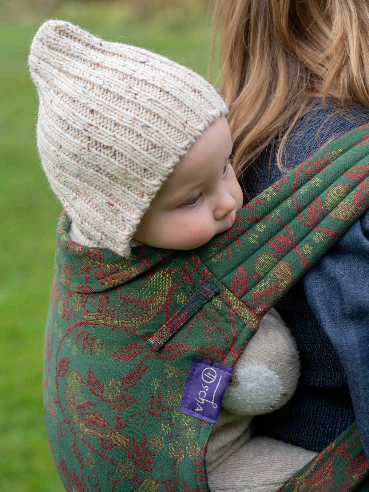 Child in a green floral-patterned carrier with a beige knit hat, outdoors.