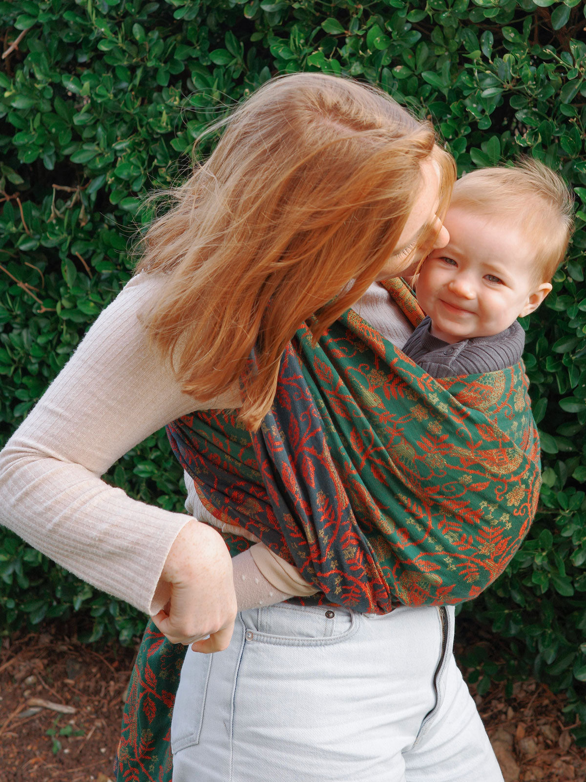 Woman carrying a baby in a green and red patterned sling against a green leafy background