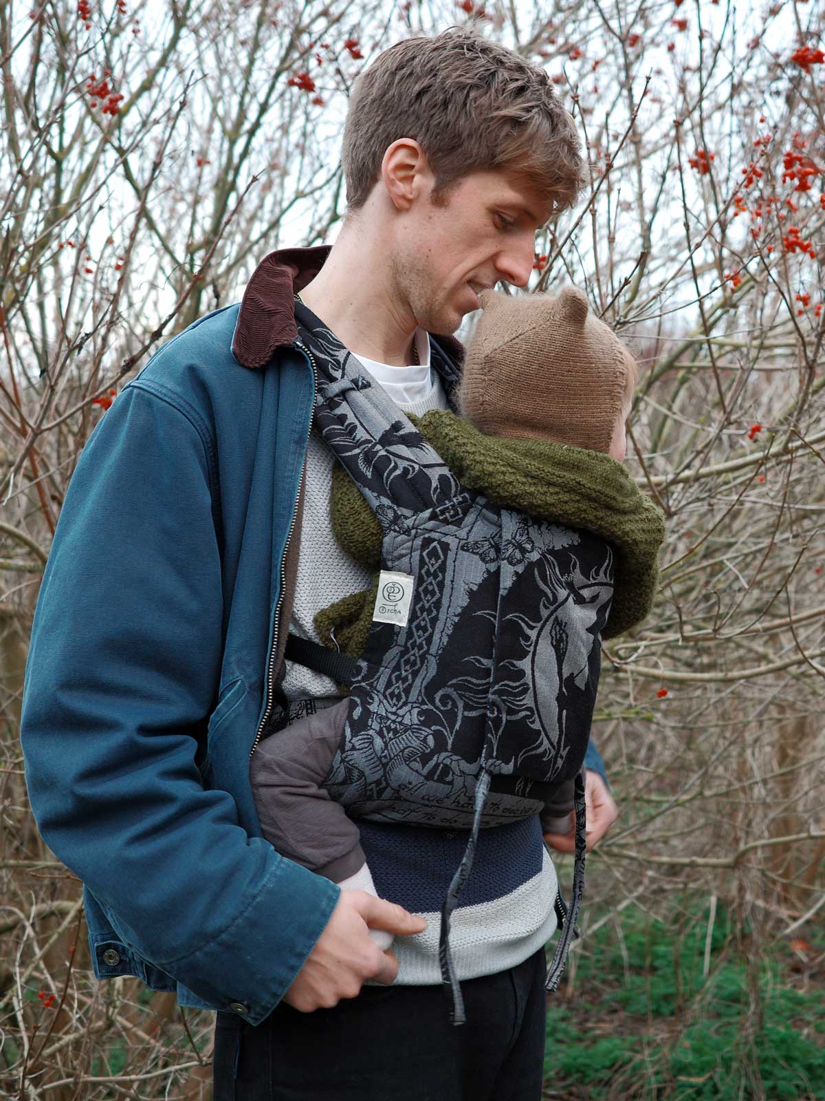 Man carrying a child in a baby carrier outdoors with bare trees and red berries in the background