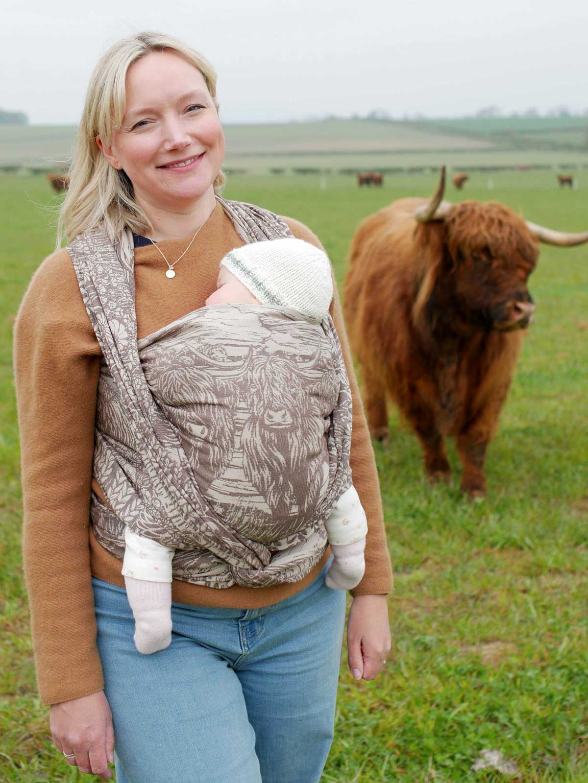 Woman holding a baby in a sling with a highland cow in the background