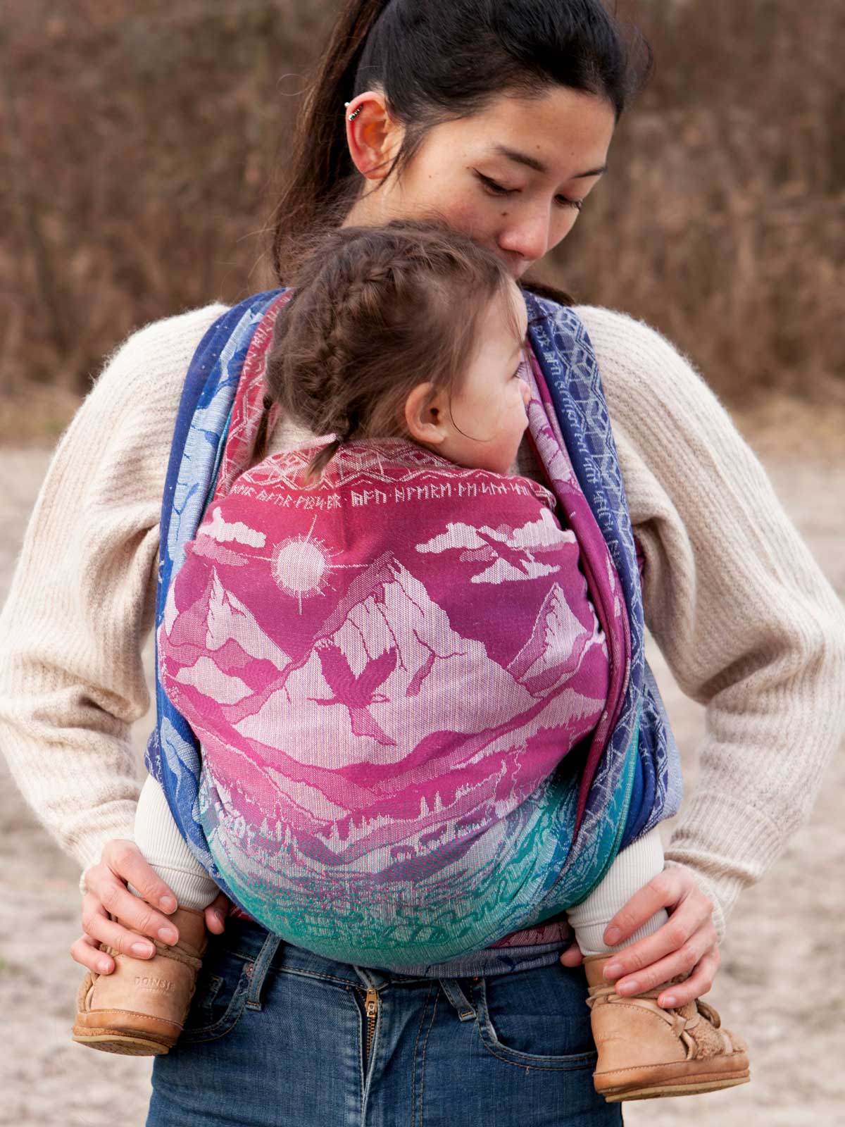 Woman carrying a child in a colorful baby carrier outdoors.