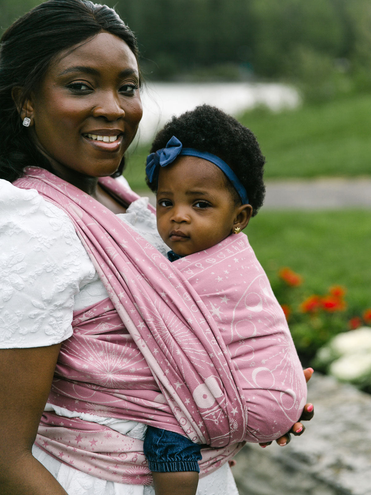 Woman holding a child in a pink sling outdoors with greenery and flowers in the background