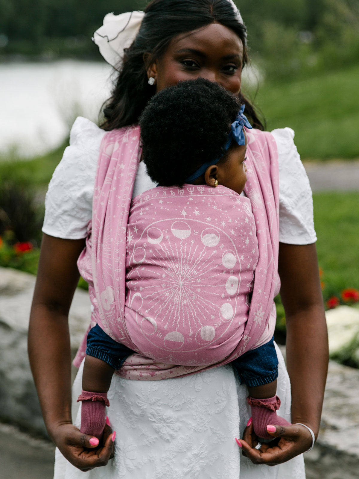 Woman carrying a child in a pink baby woven wrap outdoors near a body of water.
