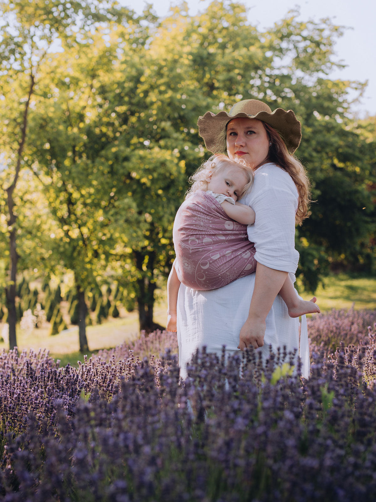 Woman holding a baby in a ring sling in a field of lavender with trees in the background