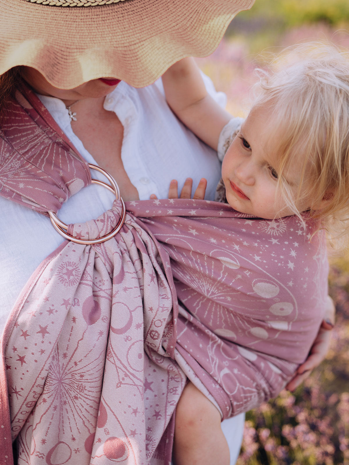 Woman holding a child wrapped in a pink ring sling with a lunar pattern, outdoors.