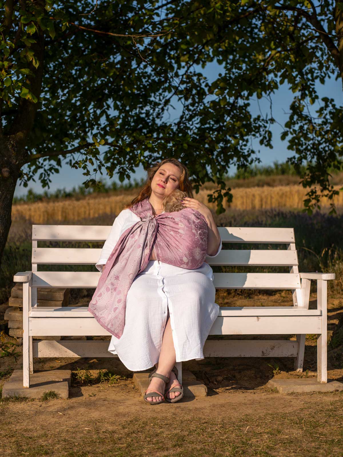 Woman sitting on a bench under a tree with a baby in a pink sling, surrounded by nature.