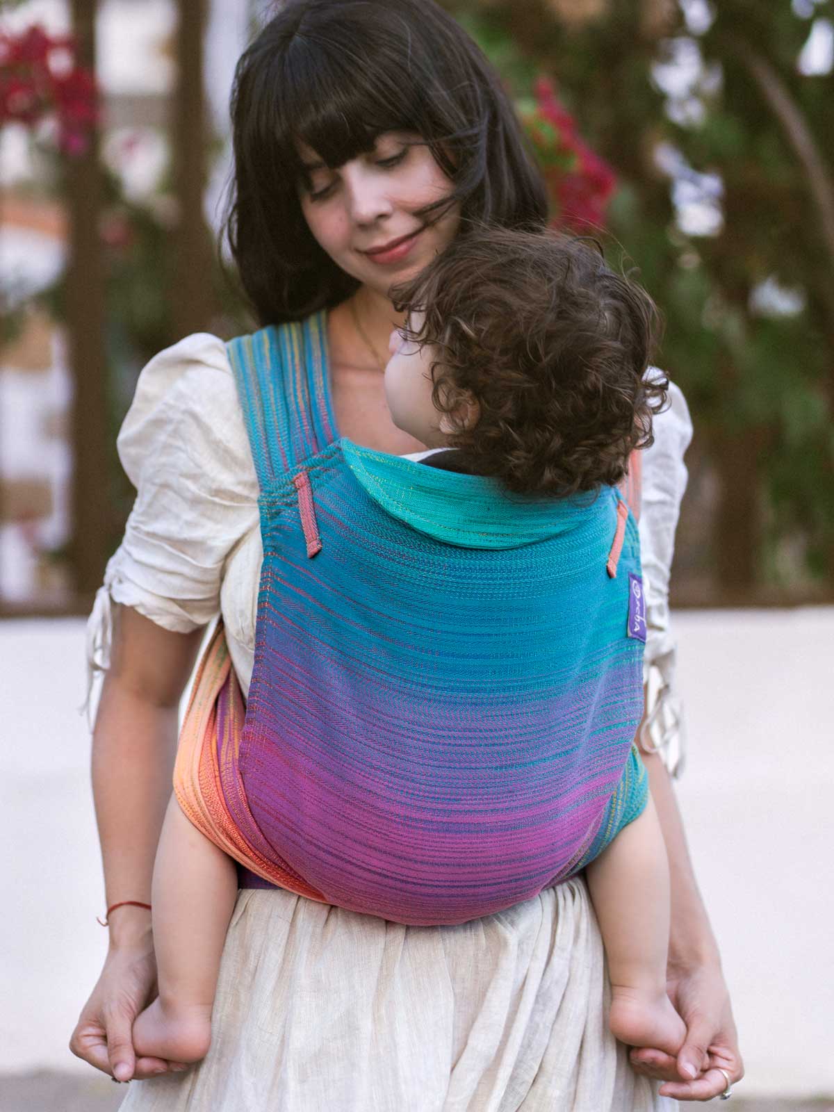 Woman carrying a child in a colorful 'Cairis' baby carrier outdoors.