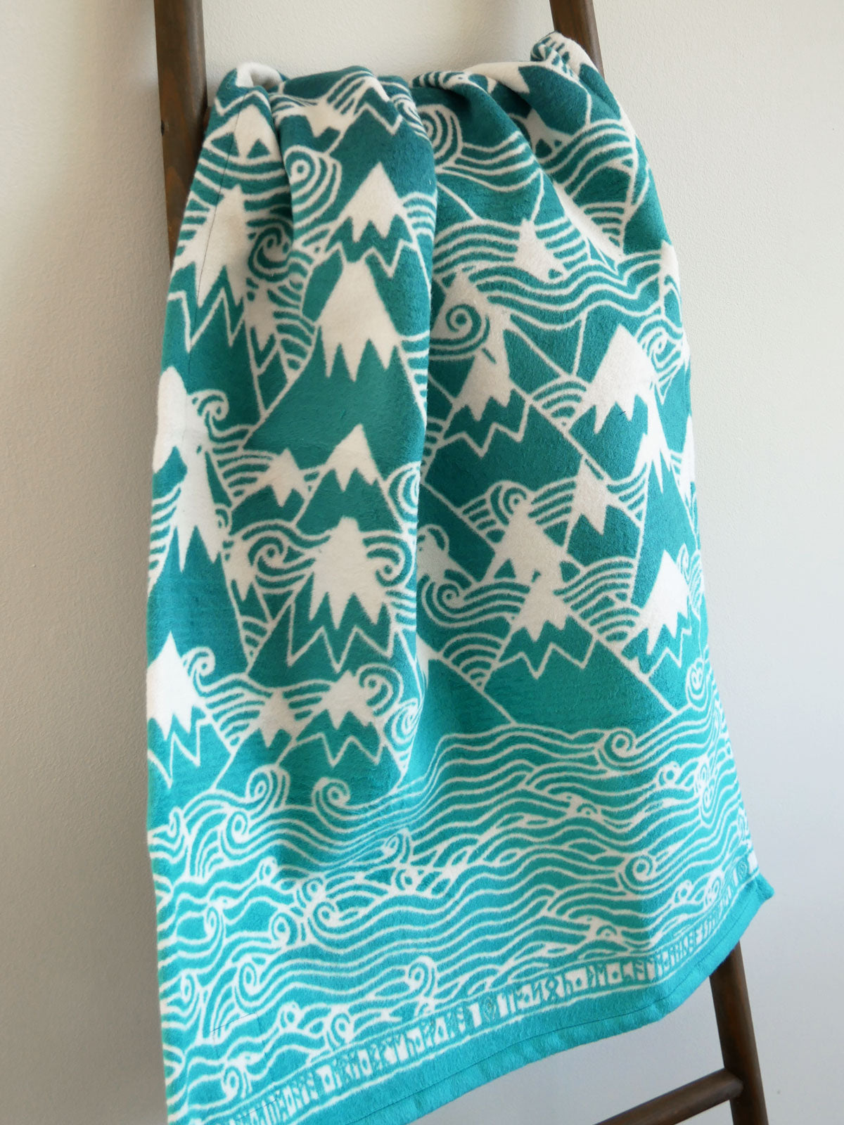 Teal baby blanket with white mountain and wave pattern draped over wooden chair.