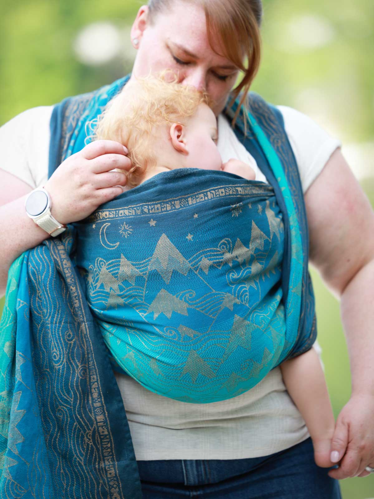 Woman carrying a baby in a blue teal woven wrap with a mountain pattern