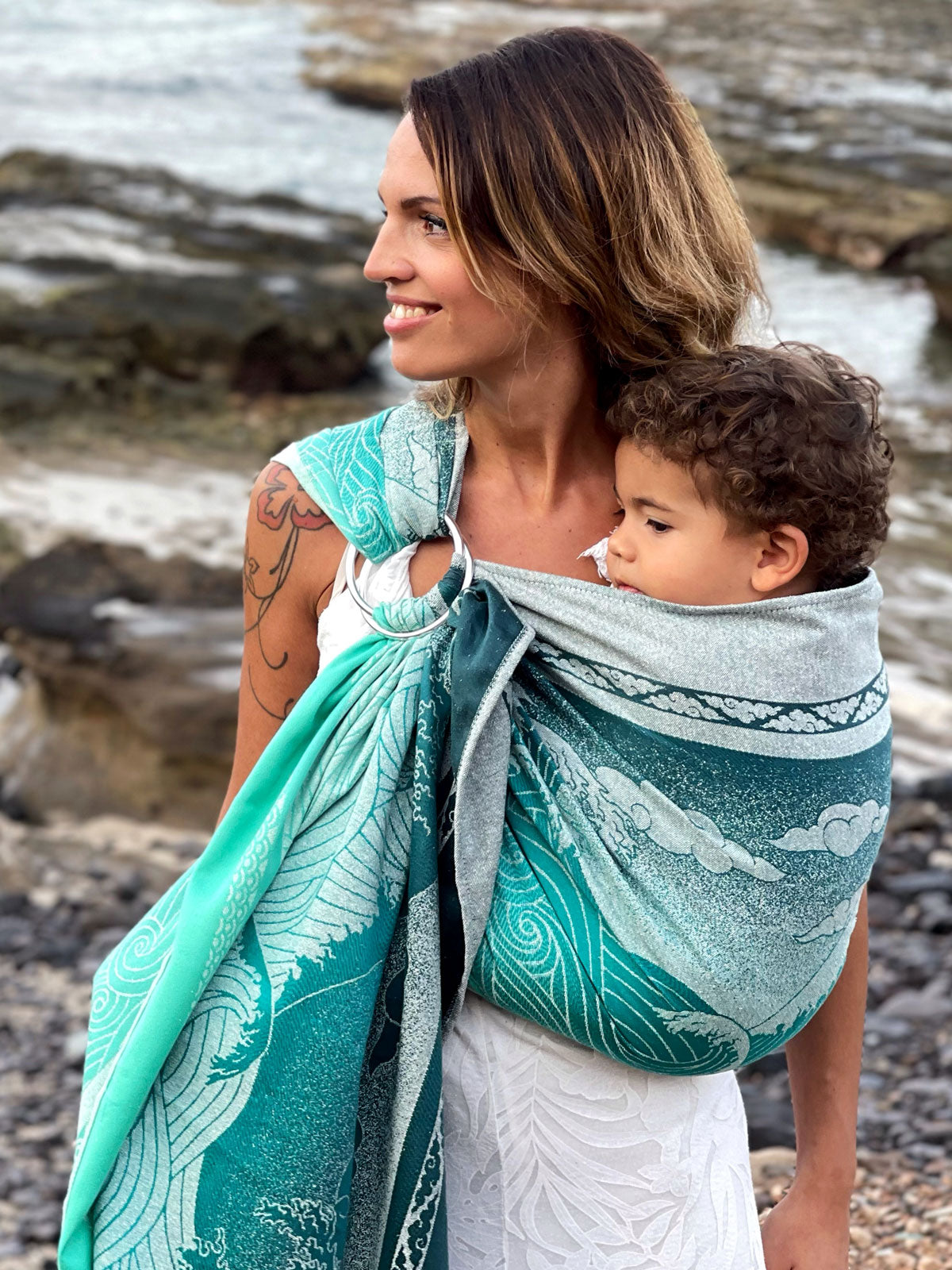 Woman holding a child in a teal and white patterned wrap on a rocky beach.