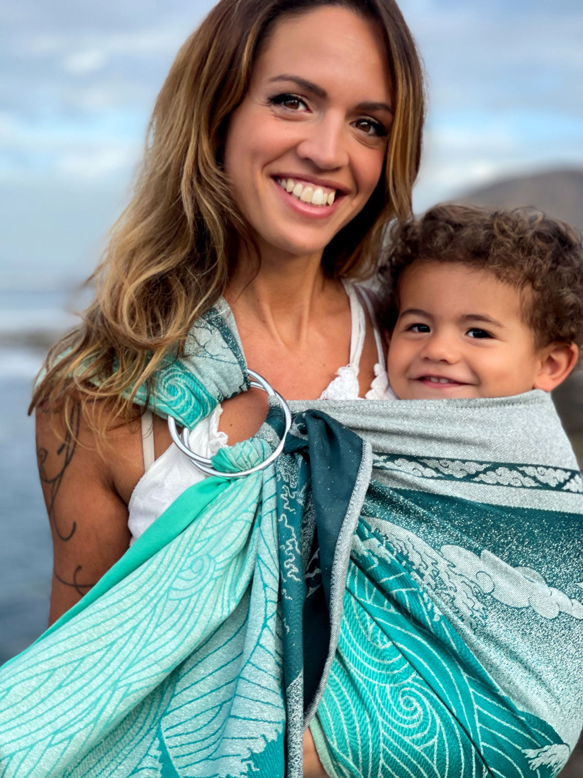 Woman holding a child in a teal and white baby carrier with a scenic background