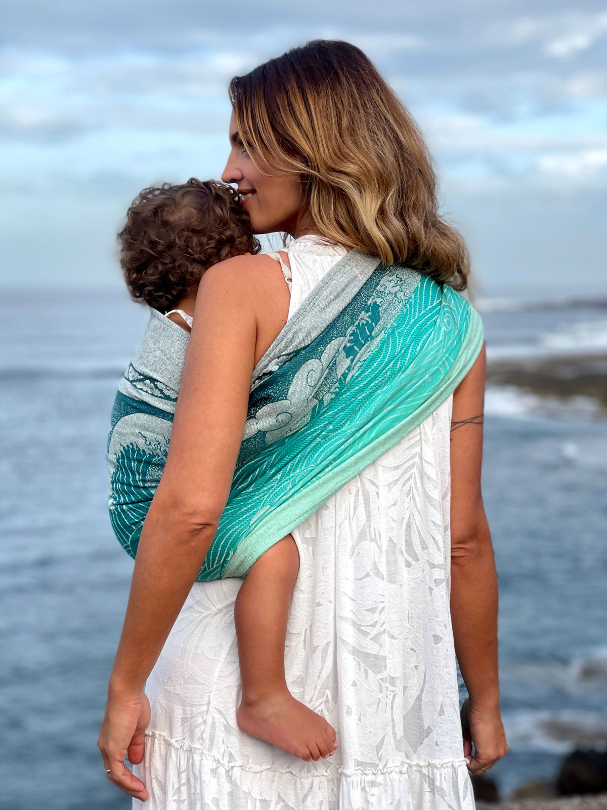 Woman carrying a child on a beach using a green and blue ring sling.