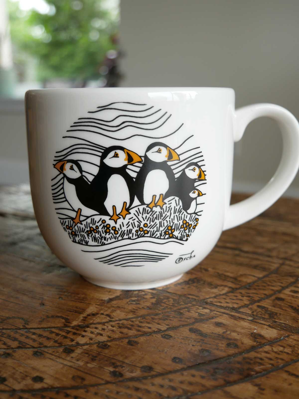 A white ceramic mug with a black and orange puffin design, sitting on a wooden surface.