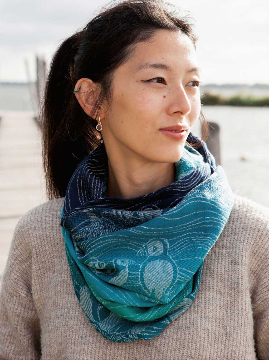 A woman standing on a pier wearing a teal and blue cowl with puffin pattern