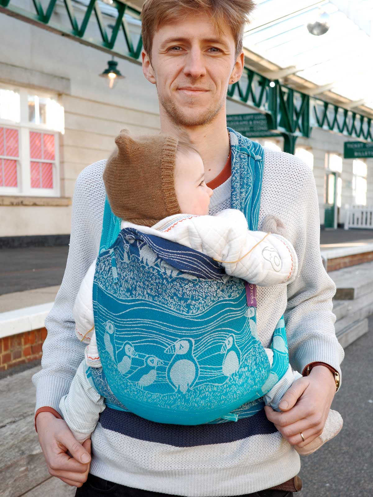 Man carrying a child in a blue baby carrier with a scenic design outdoors.