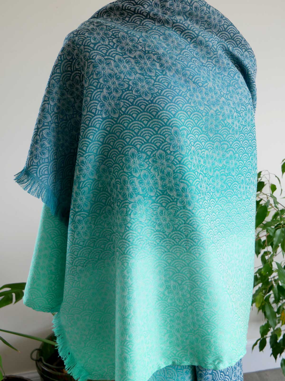 Teal and green patterned Sekaibloom Wave Scarf draped over a surface with plants in the background