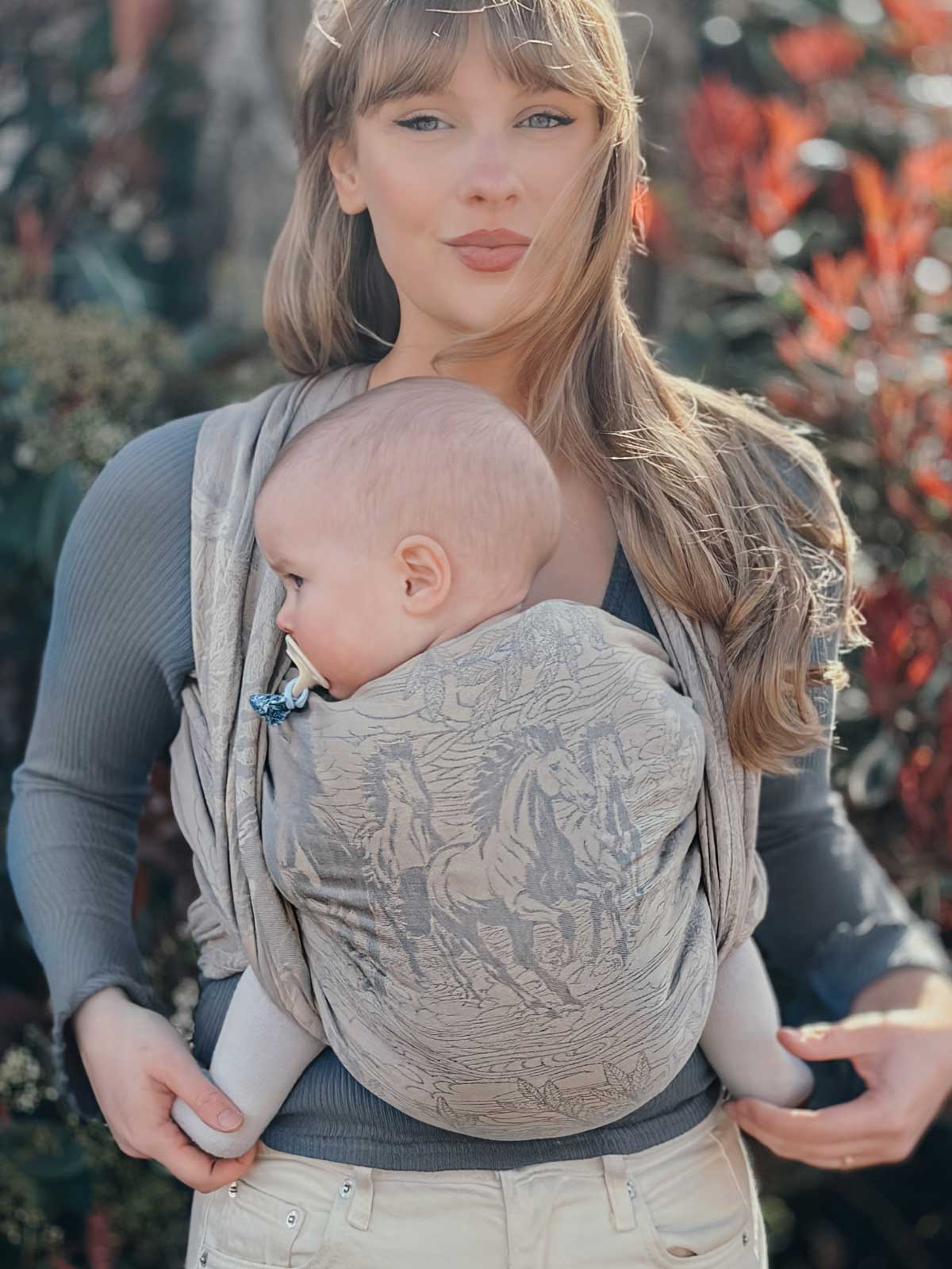 Woman holding a baby in a woven wrap featuring horses against a blurred natural background