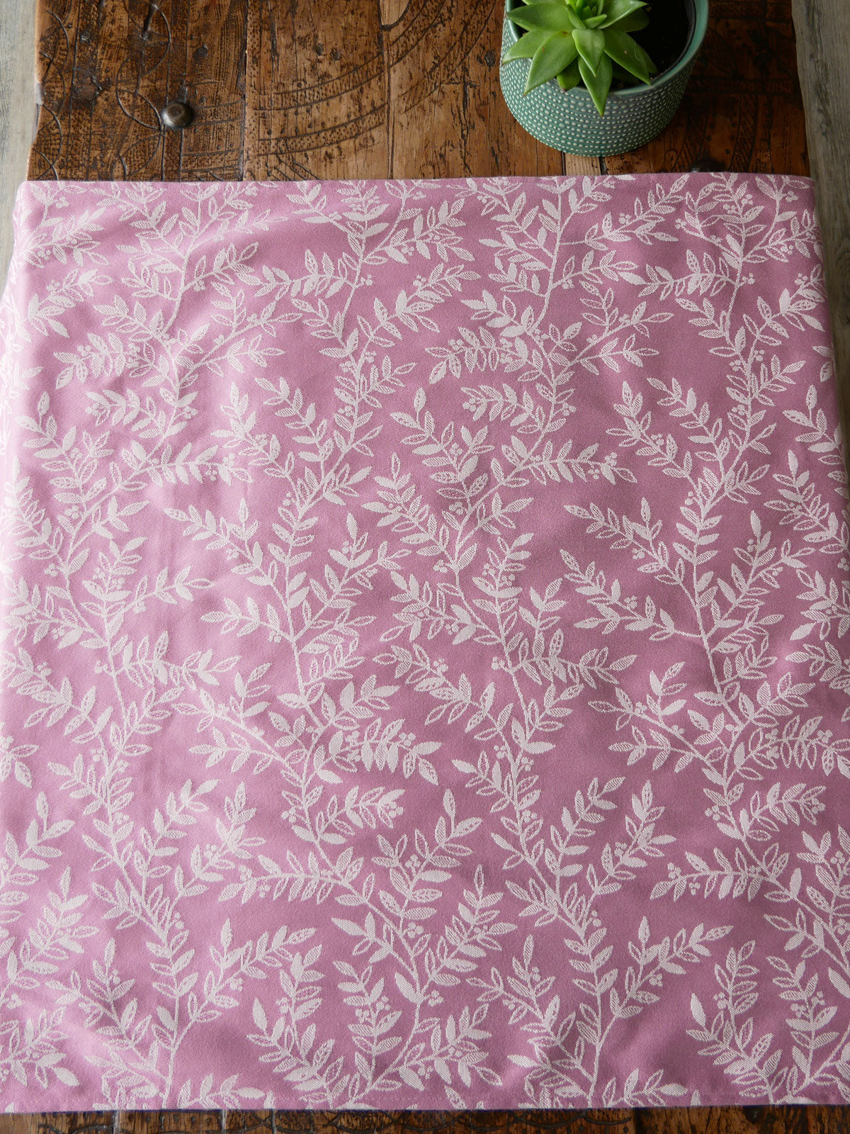 Dusky pink fabric with white leaf pattern on a wooden surface with a plant.