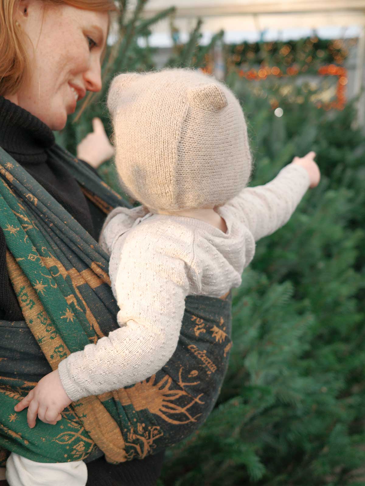 Woman holding a baby in a green and gold baby wrap with Christmas trees in the background