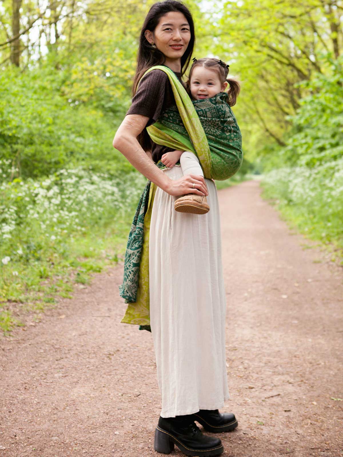 Woman carrying a baby in a green woven wrap with animal folk design.