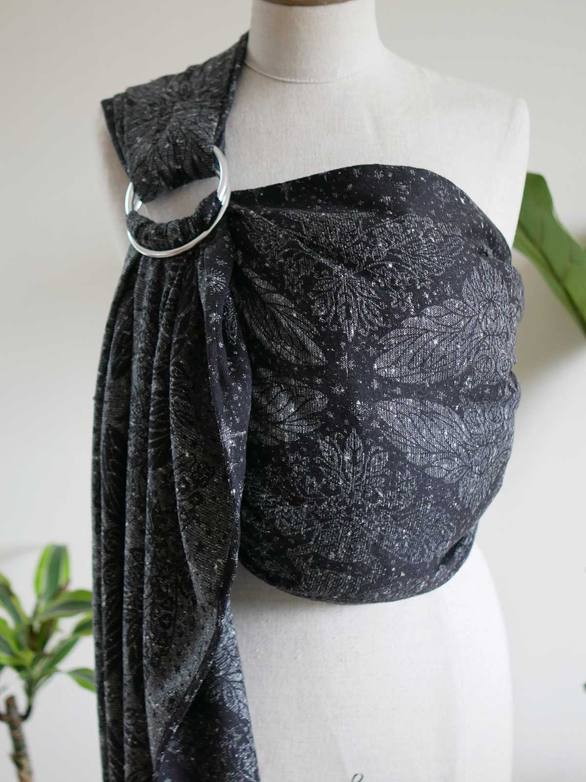 A black and gray patterned fabric baby ring sling draped on a white mannequin torso, featuring a large silver metal ring at the shoulder.