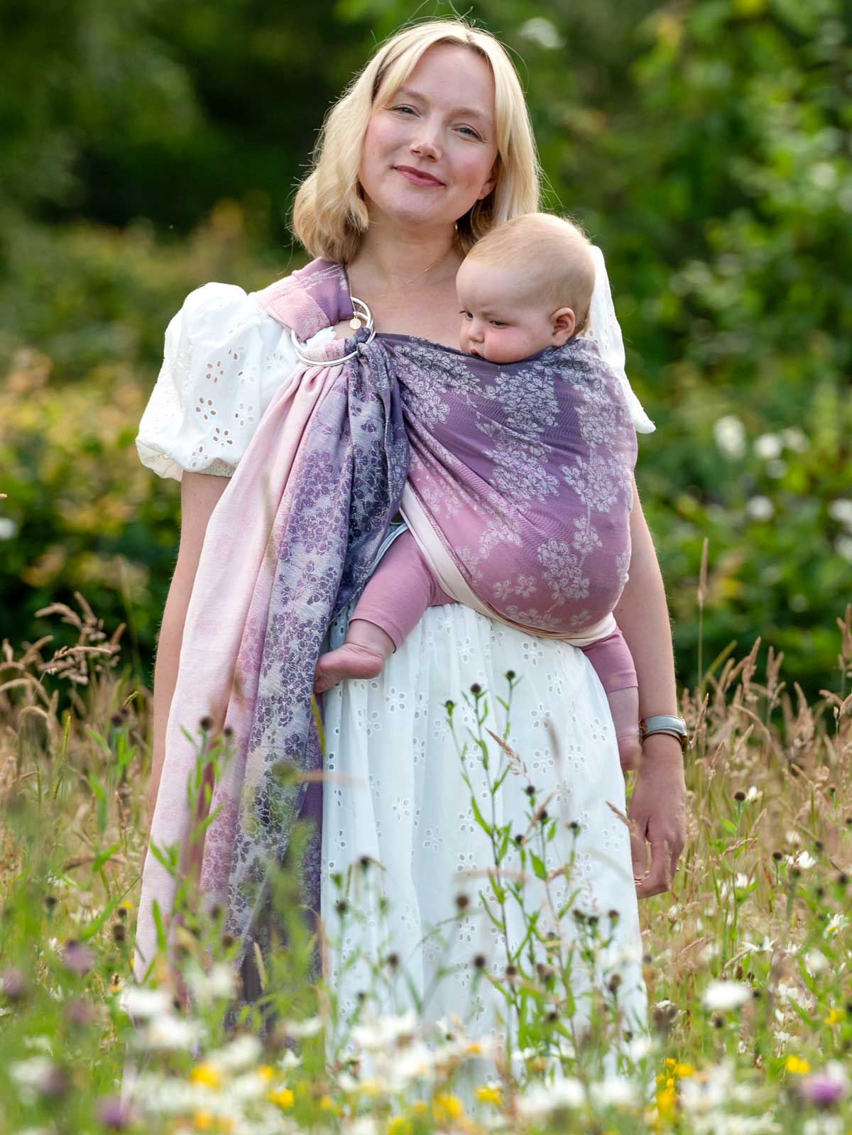 Front view of model in wild flower meadow carrying baby. 