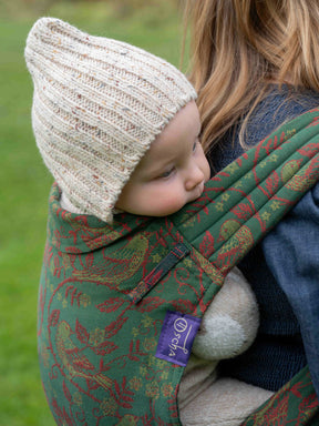 Child in a green floral-patterned carrier with a beige knit hat, outdoors.
