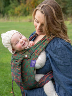 Woman holding a baby in a green baby carrier outdoors.