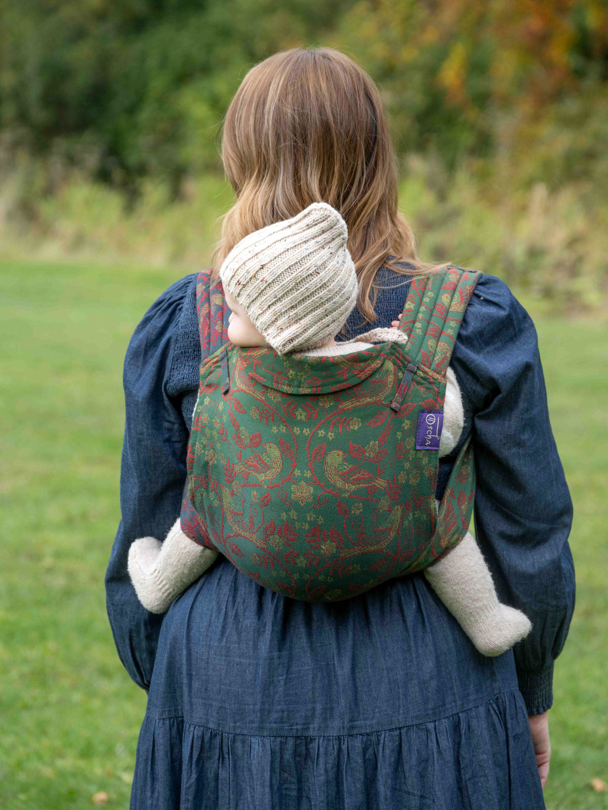 Person wearing a green baby carrier with a child inside, standing in a grassy outdoor area.
