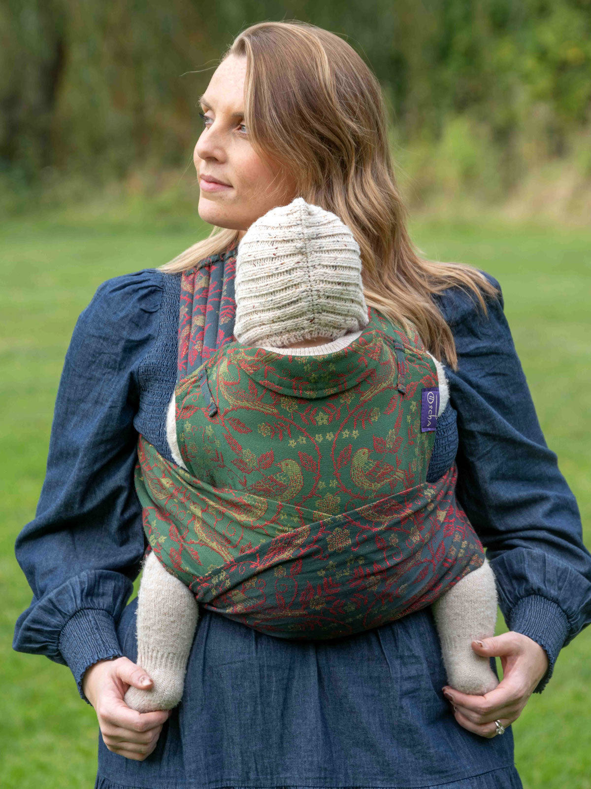 Woman carrying a child in a green and red patterned sling outdoors.