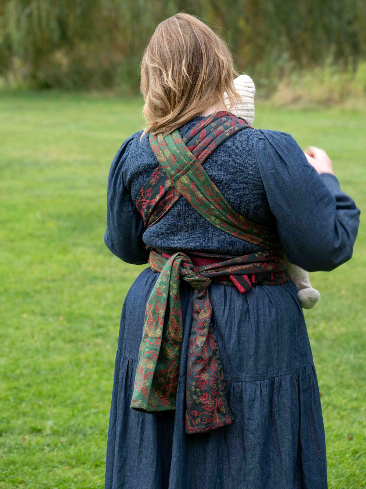 Person wearing a blue dress with a colorful baby carrier outdoors on a grassy area