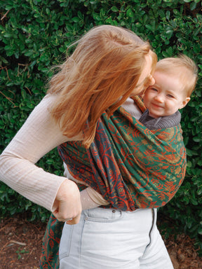 Woman carrying a baby in a green and red patterned sling against a green leafy background