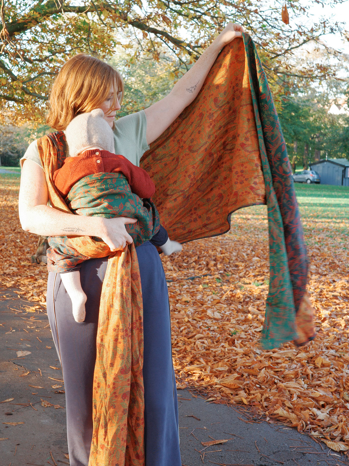 Woman holding a colorful baby wrap with a child on her back in an outdoor setting with trees and grass.