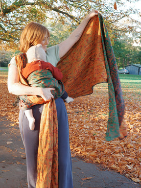 Woman holding a colorful baby wrap with a child on her back in an outdoor setting with trees and grass.