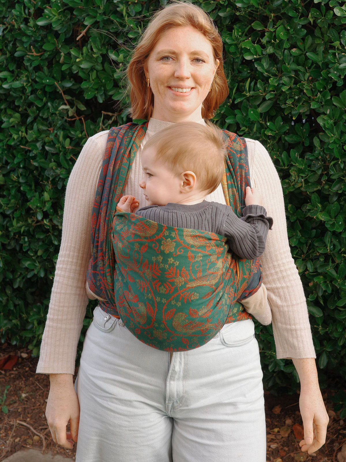 Woman carrying a baby in a floral baby wrap against a green leafy background