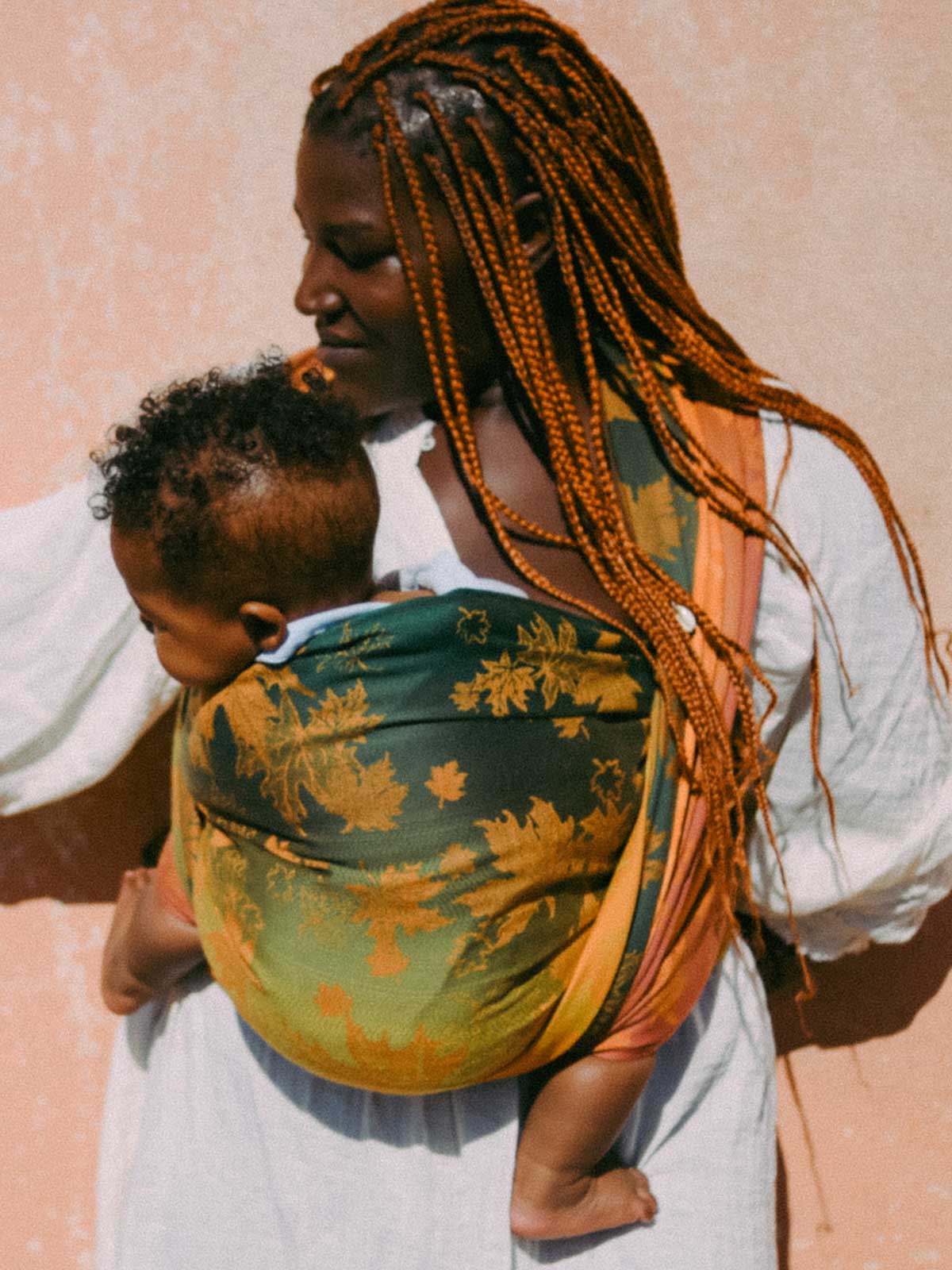 Woman holding a child in a leaf patterned baby carrier against a pink background