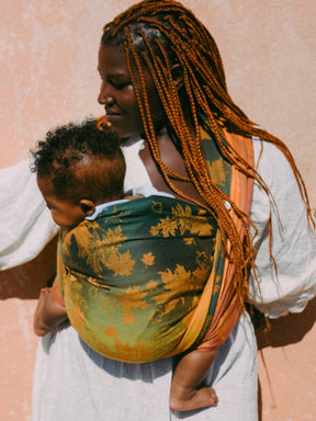 Woman holding a child in a leaf patterned baby carrier against a pink background