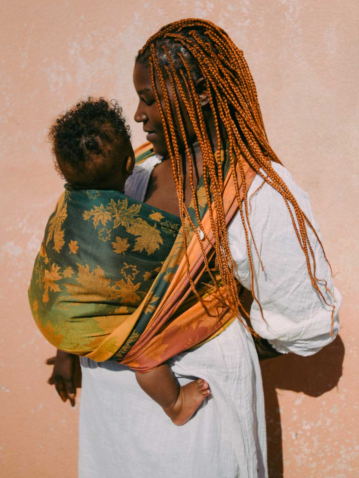 Woman holding a child wrapped in a colorful baby sling against a pink background