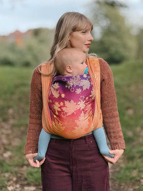 A front view of a woman carrying a baby in a autumnal coloured woven wrap with leaf patterns
