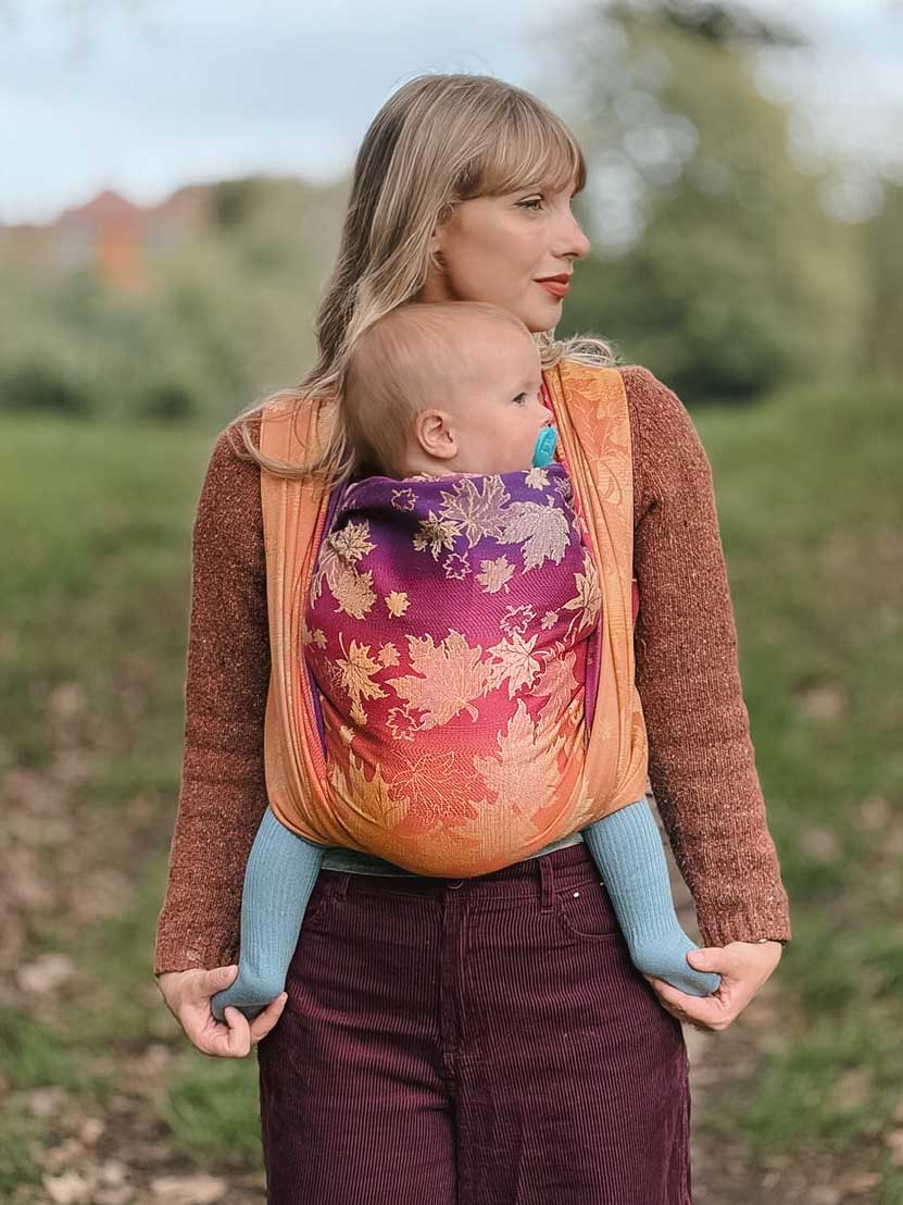 A front view of a woman carrying a baby in a autumnal coloured woven wrap with leaf patterns