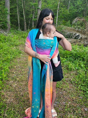 Woman holding a baby wrapped in a colorful sling outdoors in a forest setting