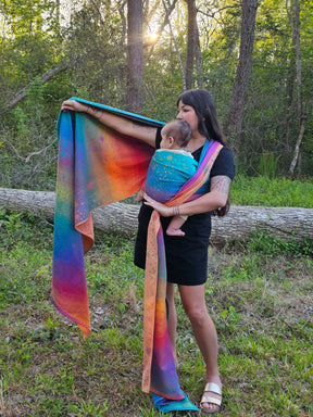 Woman holding a baby in a colorful sling outdoors with trees in the background