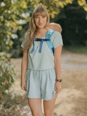 Model carries her baby on her back with shoulder and chest straps visible from the front. 