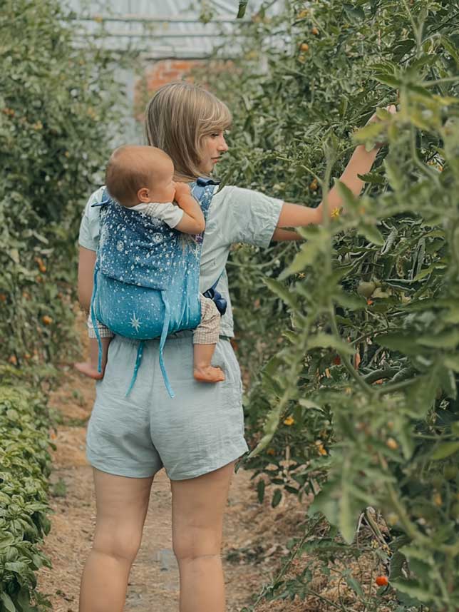 Model carries her baby in a carrier and is standing in a greenhouse full of tomato plants. 
