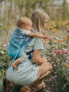 Baby being carried on Model's back. Model is crouching down in a meadow looking at flowers. 