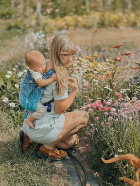 Wider view of model crouching down in a flower meadow with baby on her back. 