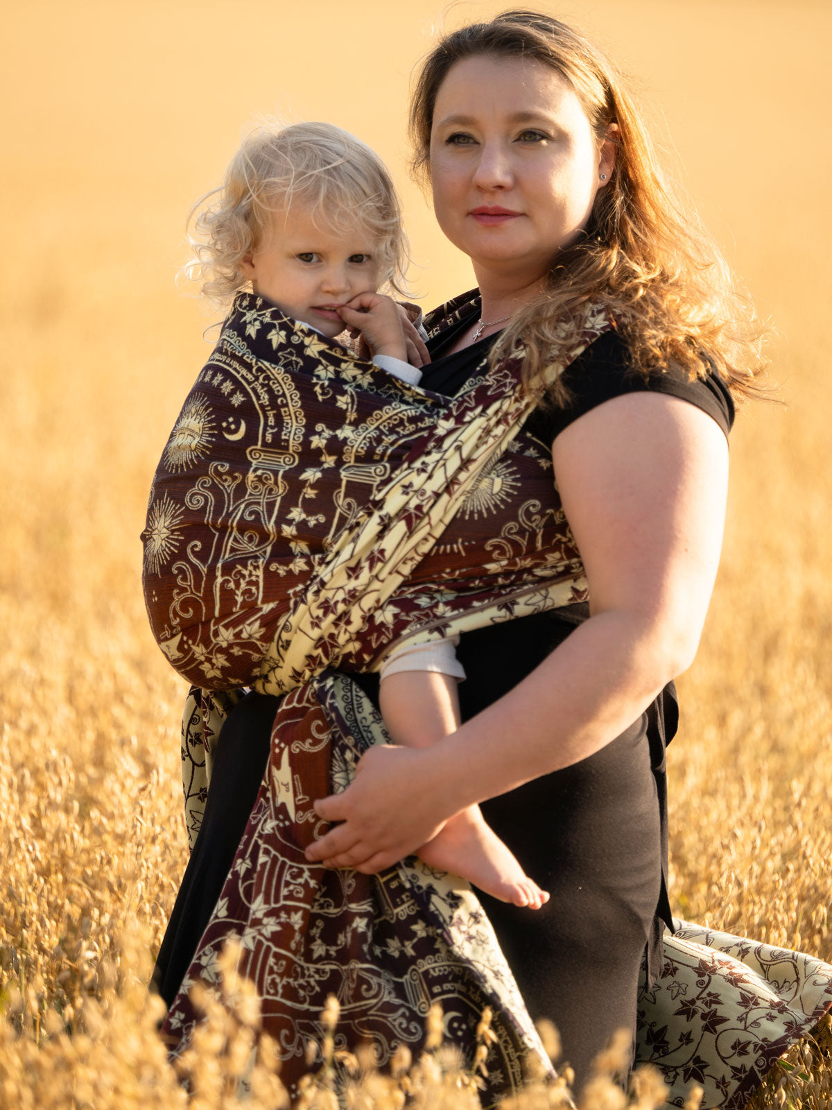 Woman holding a child in Doors of Durin Ornament Baby Wrap against a golden field background