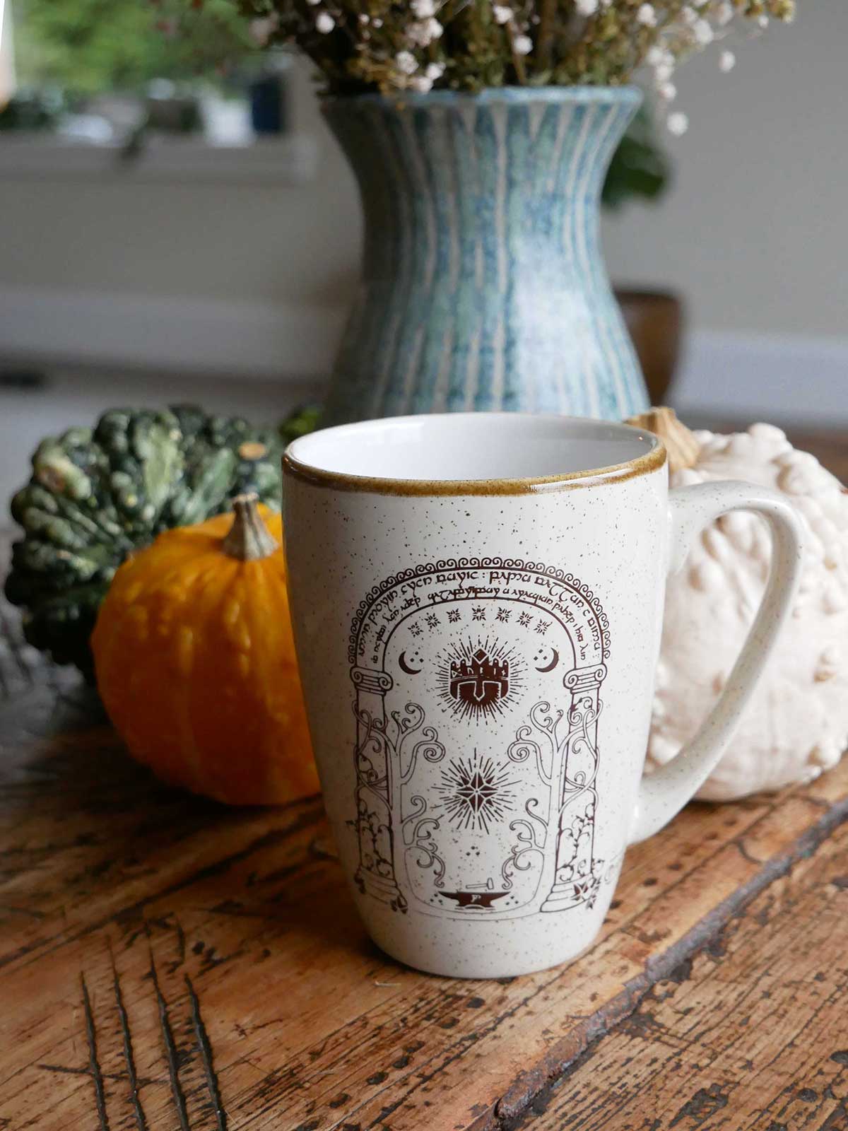 White mug with decorative design on a wooden surface with pumpkins and a vase in the background